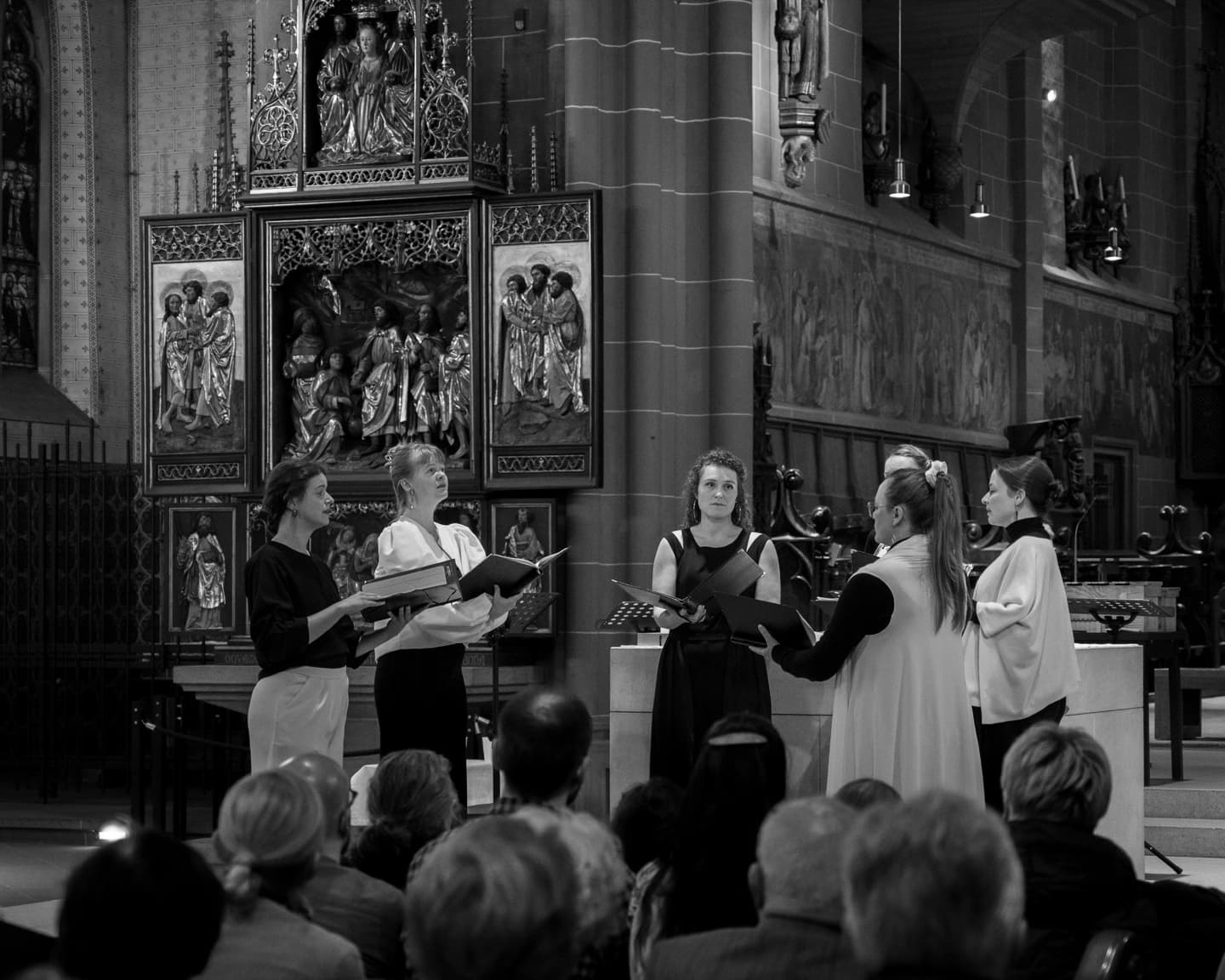Sjaella singt im Halbkreis mit Notenmappen vor einem gotischen Flügelaltar, im Vordergrund das Publikum (Schwarzweiß)