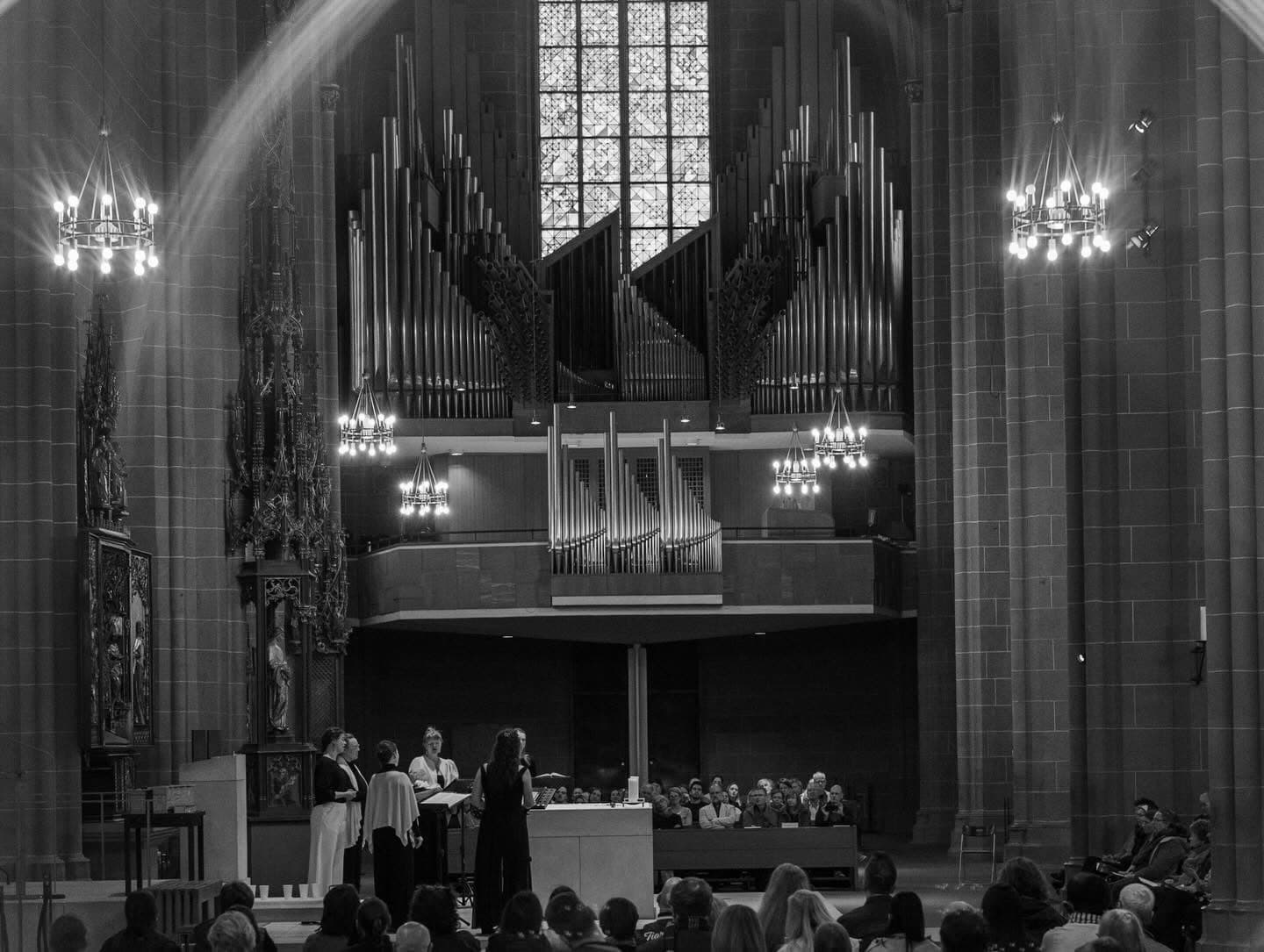 Sjaella singt im Altarraum einer gotischen Kirche vor der großen Orgel, im Vordergrund das Publikum (Schwarzweiß)